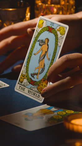 Vertical-Video-Close-Up-Of-Woman-Giving-Tarot-Card-Reading-On-Candlelit-Table-Holding-The-World-Card